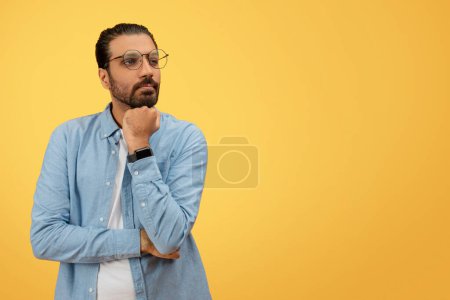 Téléchargez les photos : Un indien barbu contemplatif avec des lunettes touche son menton, portant une chemise décontractée en denim sur un fond jaune - en image libre de droit