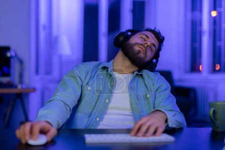 Téléchargez les photos : Un homme portant une chemise décontractée en jean et un casque s'est endormi à son ordinateur, signe d'épuisement ou de fatigue extrême. - en image libre de droit