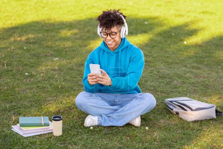 Ein junger brasilianischer Student mit Kopfhörern sitzt draußen und konzentriert sein Smartphone, umgeben von Büchern und einer Kaffeetasse