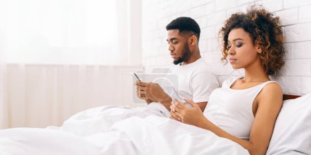 Téléchargez les photos : Jeune couple afro-américain se couche dos à dos au lit, absorbé dans leurs smartphones, illustrant les problèmes de relations modernes - en image libre de droit