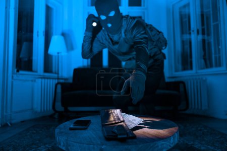 Un ladrón enmascarado en una habitación oscura usa una linterna para encontrar y robar una billetera y un teléfono en una mesa de madera