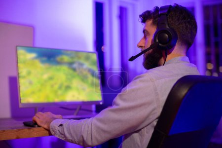 A man wearing a casual shirt with headphones intently uses a gaming computer in a room with colorful ambient lighting
