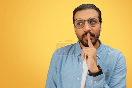Ein nachdenklicher bärtiger indischer Mann mit Brille und einem Finger auf den Lippen, der vor gelbem Hintergrund Schweigen signalisiert.