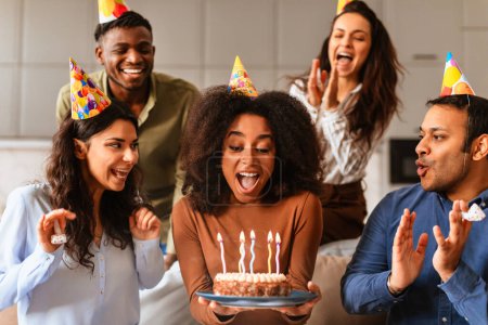 Foto de Amigos multirraciales rodean a una mujer sosteniendo un pastel de cumpleaños, soplando velas en un momento de emoción juntos - Imagen libre de derechos