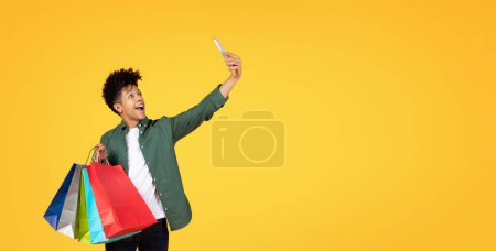 Téléchargez les photos : Jeune noir énergique capturant un moment selfie avec des sacs à provisions vibrants à la main - en image libre de droit