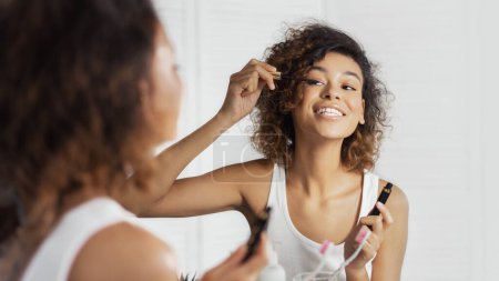 Téléchargez les photos : Afro-américaine fille mis sur mascara, faire maquillage reflétant dans miroir dans salle de bain - en image libre de droit