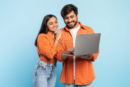 Indian couple in orange attire smiling while collaborating and looking at laptop screen, modern work concept