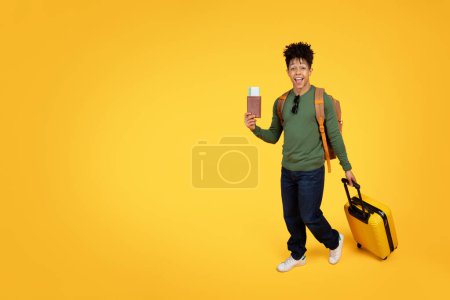 Foto de Joven y alegre afroamericano portador de pasaporte con tarjeta de embarque y tirando de una maleta amarilla, lista para viajar, sobre un fondo amarillo - Imagen libre de derechos