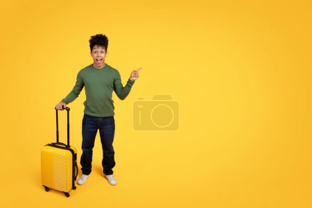 Young excited traveler african american guy pointing to side and holding a carry-on luggage on yellow backdrop