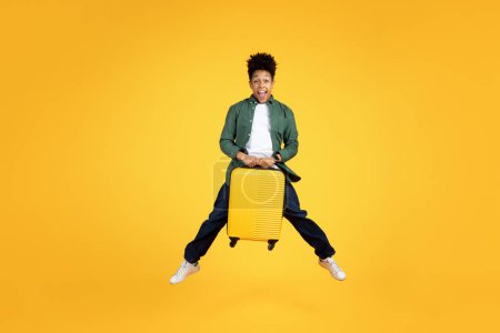Photo for An ecstatic young african american guy leaps with a bright yellow suitcase, expressing joy and the thrill of travel against a yellow backdrop - Royalty Free Image