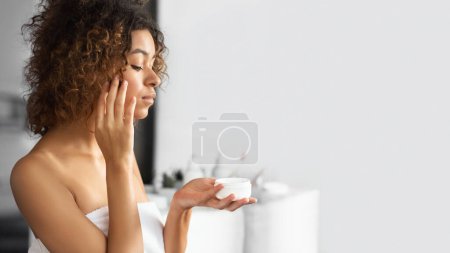 Téléchargez les photos : Jeune femme noire tenant pot de crème visage debout dans la salle de bain. Routine Soins de la peau
. - en image libre de droit