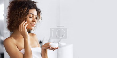 Foto de Hermosa chica afro-americana sosteniendo frasco de crema abierta, de pie en el baño
. - Imagen libre de derechos