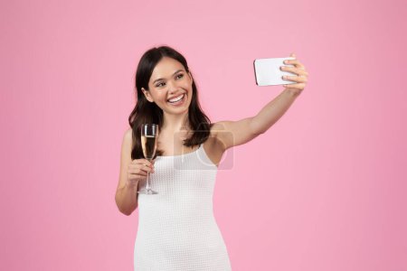 A cheerful woman in a white dress holds a champagne glass and takes a selfie with her smartphone against a pink background