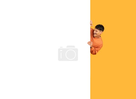 Téléchargez les photos : Un jeune homme dans un pull orange regardant ludique derrière une bannière blanche verticale vierge sur un fond orange - en image libre de droit