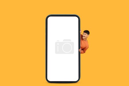 A cheerful man in an orange sweater peeking out from behind a large vertical smartphone with a blank white screen set against an orange background