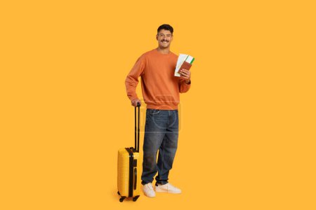 Foto de Sonriente hombre se para con maleta amarilla y tarjetas de embarque, lo que indica la preparación para un viaje contra un fondo sólido - Imagen libre de derechos