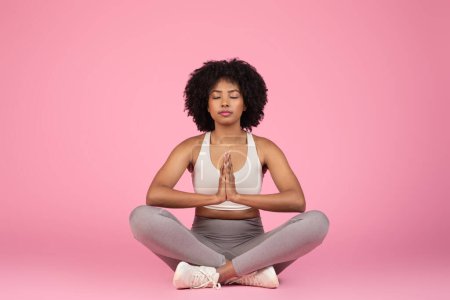 Photo for A serene African American lady sitting in a meditation pose, promoting tranquility and peace of mind, isolated on pink - Royalty Free Image