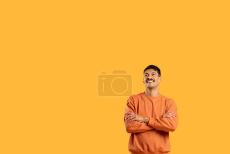 Photo for A happy, confident man in a casual orange sweater stands with arms crossed against a solid yellow background, looking upwards with a smile - Royalty Free Image