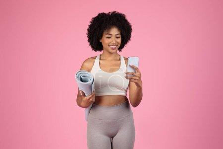 Foto de Mujer deportiva afroamericana sosteniendo una esterilla de yoga y mirando un teléfono inteligente, lo que sugiere un estilo de vida activo moderno, aislado en rosa - Imagen libre de derechos