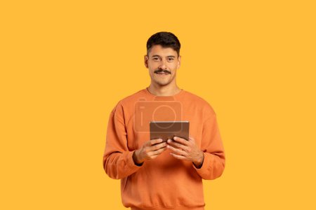 Fröhlicher Mann in orangefarbenem Pullover hält ein Tablet mit weichem Lächeln, orangefarbenem Hintergrund, surft oder scrollt