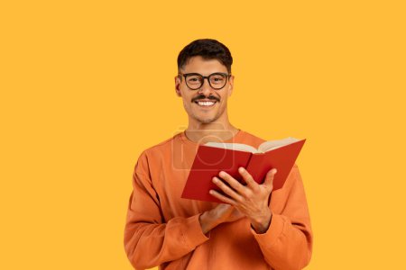 Young smiling man with glasses enjoying a red book on a vibrant orange backdrop, leisure and education concept