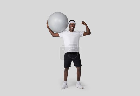 Téléchargez les photos : Un Afro-Américain en forme sourit en soulevant une grosse balle d'exercice au-dessus de sa tête dans un étalage de force et de remise en forme sur un fond gris - en image libre de droit