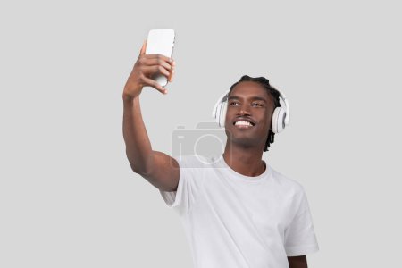 Un homme afro-américain joyeux portant des écouteurs blancs et un t-shirt blanc prend un selfie avec son smartphone sur un fond gris