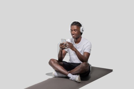 Foto de Un joven afroamericano con una camiseta blanca y auriculares está sentado con las piernas cruzadas en una alfombra, sosteniendo y mirando un teléfono inteligente con una expresión alegre - Imagen libre de derechos