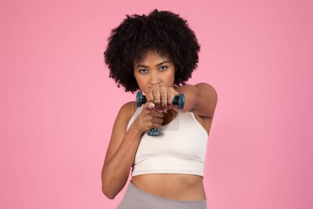 Determined african american lady performing bicep curls with a dumbbell, emphasizing strength and focus, isolated on pink