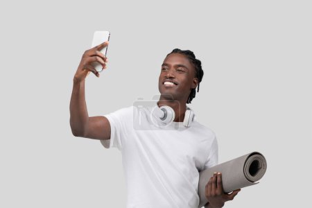 A happy young African American man with dreadlocks is holding a smartphone to take a selfie while carrying a yoga mat and wearing headphones