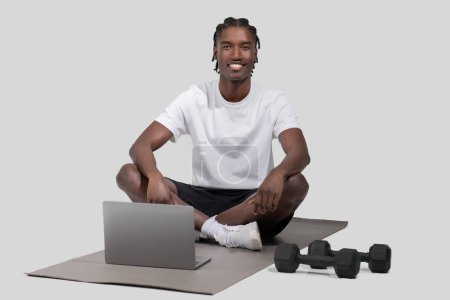 A relaxed african american guy sits on a gym mat with a laptop and weights beside him, in a casual setting isolated on a white backdrop