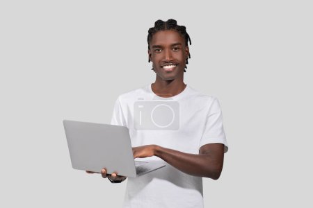 A photo depicting a smiling black guy holding a laptop computer, posing casually and looking at camera isolated on white