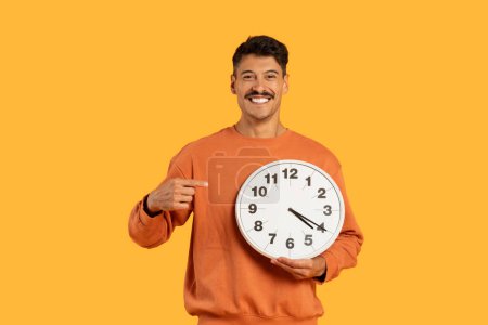 A millennial guy with a moustache holds a large clock, showcasing punctuality and time management in a funny way, isolated on yellow