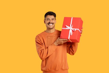 Photo for Man in orange sweater seemingly offering a red gift box with a bright smile and a sense of anticipation - Royalty Free Image