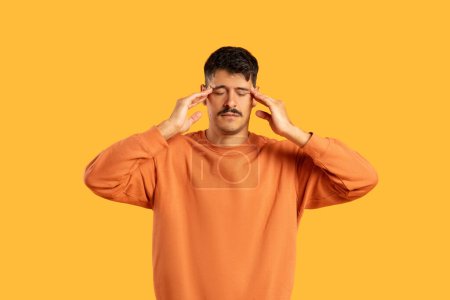 Homme avec moustache en pull orange tenant la tête, montrant des signes de maux de tête ou de stress sur fond jaune