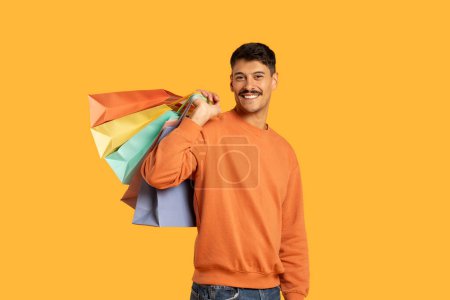 Foto de Un tipo milenario con un bigote que lleva coloridas bolsas de compras sobre un fondo amarillo aislado, con aspecto divertido y contenido - Imagen libre de derechos