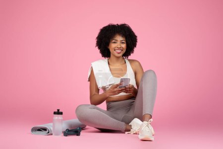 Téléchargez les photos : Une Afro-Américaine détendue assise les jambes croisées tenant un smartphone, avec des essentiels de gym autour, isolée sur rose - en image libre de droit