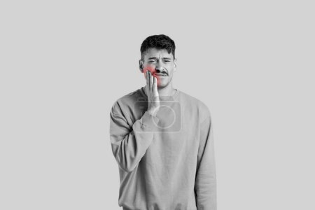 Téléchargez les photos : Une image en noir et blanc montre un homme exprimant des maux de dents en se frottant la joue, indiquant potentiellement des douleurs à la mâchoire, sur un fond gris uni - en image libre de droit