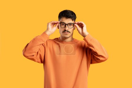Un homme réfléchi avec moustache en pull orange ajuste ses lunettes sur un fond jaune vif