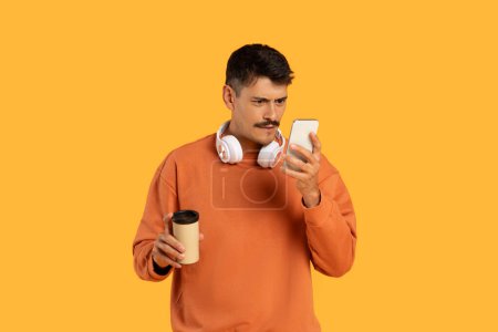 A millennial guy with moustache examines smartphone, funny and focused, with coffee, isolated on orange