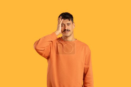 A young man wearing an orange sweater is posing with his hand on his forehead, indicating a headache or frustration, against an orange background