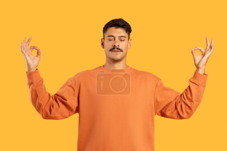 Photo for Calm man with moustache meditating with eyes closed and gesturing Zen peace in a serene pose on a yellow backdrop - Royalty Free Image