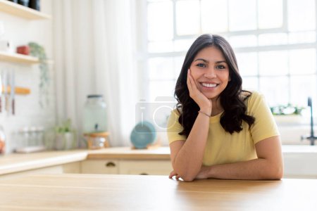 Porträt einer lächelnden Frau aus dem Mittleren Osten, die auf einem Küchentisch mit hellem, einladendem Hintergrund lehnt, Kopierraum