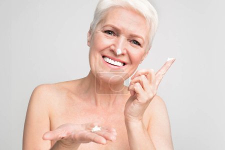 Lebendige ältere europäische Frau lächelt und präsentiert eine Vielzahl von Hautpflegecremes, die s3niorlife und Wohlbefinden verkörpern