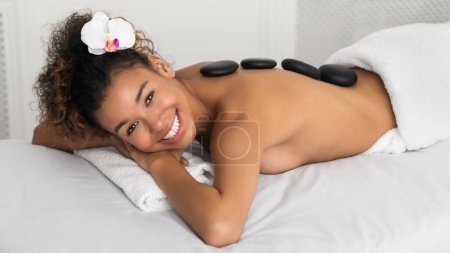 Foto de Mujer afroamericana feliz disfrutando de masaje de piedra caliente en el salón de spa - Imagen libre de derechos
