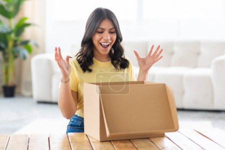 An excited middle eastern young woman in a yellow shirt is gleefully opening a brown cardboard box at home