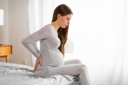 Foto de Una joven embarazada con una camisa a rayas parece estar experimentando dolor en la parte baja de la espalda mientras sostiene su espalda con una expresión dolorosa en un dormitorio bien iluminado - Imagen libre de derechos