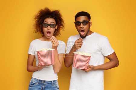 Amazed black man and woman in 3d glasses enjoying movie and eating popcorn, orange studio background