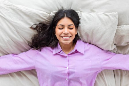 A middle eastern woman wearing a purple shirt lies on her bed with her arms outstretched, exuding a feeling of freedom and relaxation