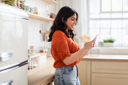 Foto de Una mujer atractiva casualmente apoyada en un mostrador de cocina mientras está ocupada con su teléfono inteligente, buscando una buena receta - Imagen libre de derechos